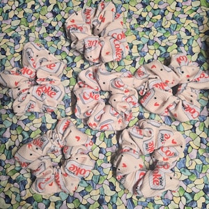 May include: Five fabric hair scrunchies in a light beige color featuring a repeating pattern of Diet Coke soda cans, small red hearts, and tiny black stars. The scrunchies have a gathered, ruffled texture and are arranged in a group.