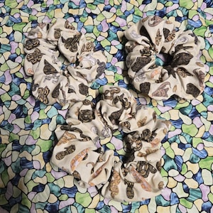 May include: Three fabric hair scrunchies featuring a repeating pattern of various snakes in shades of brown, tan, and black on a cream-colored background. The scrunchies are circular with gathered, ruffled fabric edges and a flexible elastic interior.