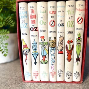 May include: A set of seven vintage Oz book series by L. Frank Baum, in a red box. The book spines are white with colorful illustrations of characters like the Scarecrow, Tin Woodman, and Wizard. The titles are in black and green.
