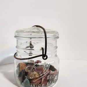 May include: A clear glass Ball jar with a lid and wire handle, filled with colorful stones. The jar has embossed text and a wide mouth design. The stones are various colors, including brown, green, and red.