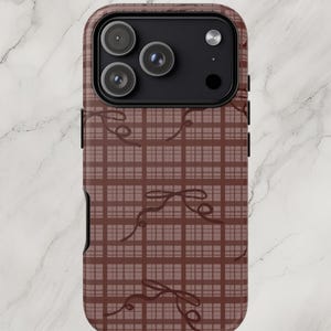 May include: A brown phone case featuring a plaid pattern with delicate bow accents. The case has a glossy finish and a camera cutout for a smartphone. The design is repeated across the surface.