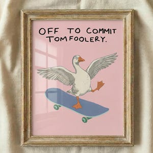 May include: Framed art print showing a goose on a skateboard with its wings spread, set against a pink backdrop. The text "OFF TO COMMIT TOMFOOLERY" is at the top. The frame has a distressed, light brown finish.