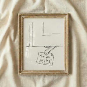 Könnte beinhalten: Eine gerahmte Bleistiftskizze zeigt eine Katzenpfote, die einen Zettel mit der Aufschrift "Are you pooping? I miss you :(" hält. Das Kunstwerk befindet sich in einem rustikalen, hellen Holzrahmen vor einem cremefarbenen Stoffhintergrund.