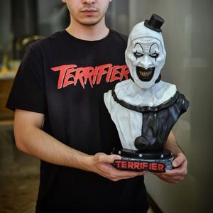 Könnte beinhalten: Ein schwarzes T-Shirt mit dem roten Schriftzug Terrifier und eine weiß-schwarze Büste der Figur Art the Clown mit einem kleinen schwarzen Zylinder, einem breiten Grinsen mit dunklen Zähnen und einem Sockel mit dem roten Schriftzug Terrifier.