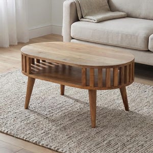 Könnte beinhalten: Ovaler Couchtisch aus hellem Holz mit Lamellendesign an den Seiten und einem unteren Regal. Der Tisch hat vier abgewinkelte Beine und ist aus hellem Holz gefertigt. Er steht auf einem neutral getönten Teppich in einem Wohnzimmer.