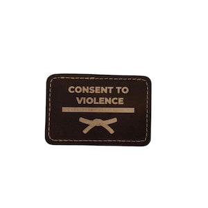 Könnte beinhalten: Ein rechteckiger, dunkelbrauner Aufnäher mit weißen Nähten. Der Text "CONSENT TO VIOLENCE" ist weiß gedruckt, über einer horizontalen Linie und einer stilisierten Schleife.