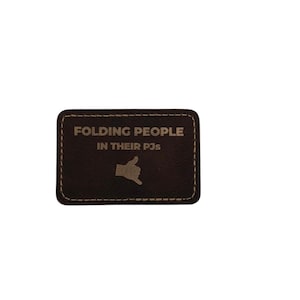 May include: A rectangular dark brown leather patch with rounded corners and light tan stitching along the border. The text FOLDING PEOPLE IN THEIR PJs is engraved in the center above a small graphic of a hand making a shaka sign gesture.