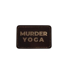 May include: A rectangular dark brown leather patch with rounded corners and visible stitching along the edges. The text MURDER YOGA is embossed in a light tan color in the center of the patch in a bold, sans-serif capital letter font.