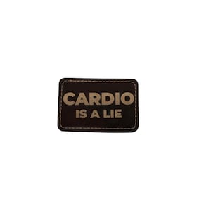 May include: A rectangular, dark brown patch with the words "CARDIO IS A LIE" in a light brown, bold font. The patch has a stitched border and a slightly distressed appearance, suggesting a leather or similar material.