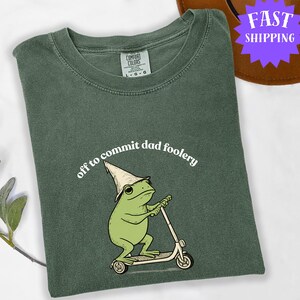 May include: Green Comfort Colors t-shirt featuring a cartoon frog riding a scooter and wearing a hat. The text "off to commit dad foolery" is arched above the frog. The shirt has a crew neck and a relaxed fit.