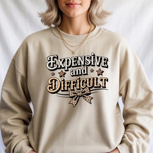 Puede incluir: Sudadera beige con el texto "Expensive and Difficult" en una fuente retro. El diseño incluye estrellas y un lazo con estampado de leopardo. La sudadera es de color neutro y tiene cuello redondo.