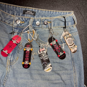 May include: Blue denim jeans featuring a Social Collision brand label at the waist. Attached to the belt loops are multiple silver-toned carabiners and chains holding five miniature finger skateboards in various colors, including red, black, and white.