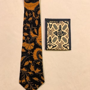 May include: A black tie and matching pocket square with a floral and leaf pattern in shades of orange and gold. The pocket square has a dark blue border and the text "Batik Keris".