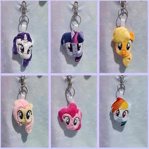 May include: Six plush keyrings featuring cartoon pony faces in various colours. Each keyring has a silver-tone clip and a white strap. The ponies include purple, blue, yellow, pink, and rainbow manes and features.