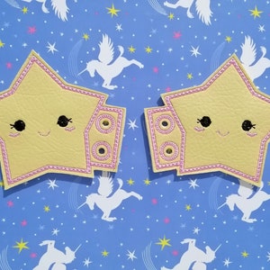 Puede incluir: Dos parches amarillos en forma de estrella con ribete rosa y caras bordadas. Cada parche tiene una extensión rectangular con detalles circulares. El fondo es azul con patrones de estrellas y unicornios blancos. Estos parches son probablemente para manualidades.