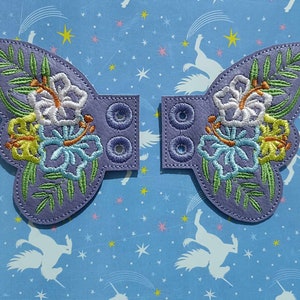 May include: Two lavender-colored butterfly wing patches with embroidered floral designs. Each wing features a cluster of white, yellow, and blue flowers with green leaves. The patches have a rectangular section with four embroidered holes.