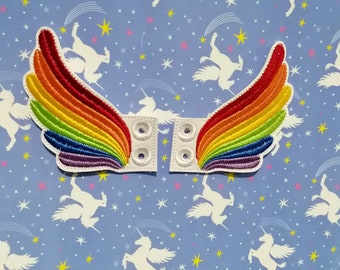 Puede incluir: Dos alas bordadas con arcoíris con respaldo blanco y cuatro ojales. Las alas son de los colores del arcoíris, con rojo, naranja, amarillo, verde, azul y morado. El fondo es una tela azul claro con unicornios blancos y estrellas amarillas.