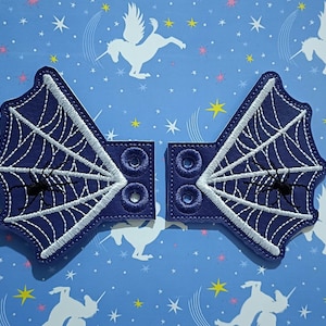 Op de afbeelding: Twee donkerblauwe, geborduurde patches met een spinnenwebontwerp. Elke patch heeft een wit web, een zwarte spin en een vierkante basis met twee decoratieve cirkels. De patches zijn ontworpen voor een Halloween- of gothic-thema.