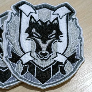 Peut inclure: Écusson brodé représentant une tête de loup dans un écu. Le patch est réalisé avec des fils noirs, blancs et gris, avec des coutures détaillées. Le loup est centré, entouré d'éléments décoratifs.