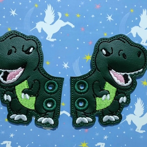 Puede incluir: Dos parches de dinosaurio verde oscuro con vientres verde claro y bocas rosas. Los parches tienen un diseño cosido y están sobre un fondo azul con patrones de estrellas y unicornios blancos. Cada parche tiene dos juegos de agujeros.