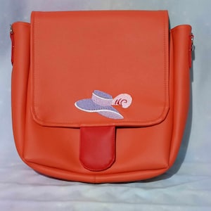 May include: A bright orange crossbody bag with a flap and red detail. It has an embroidered hat and feather design. The bag has a shoulder strap and is made of a smooth material. The bag is suitable for everyday use.
