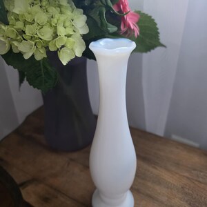 Pode incluir: Um vaso de vidro branco opalescente com uma borda alargada e uma base arredondada. O vaso é esguio e elegante, com uma superfície lisa. Um buquê de flores coloridas, incluindo flores roxas, rosas e verdes, está ao fundo.