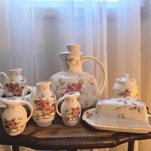 Könnte beinhalten: Eine Sammlung cremefarbener Keramik-Servierteile mit floralem Design. Das Set umfasst einen großen Krug, kleine Krüge, eine Butterdose, eine Zuckerdose mit Deckel und eine Servierschüssel, alle mit rosa und braunen floralen Akzenten.