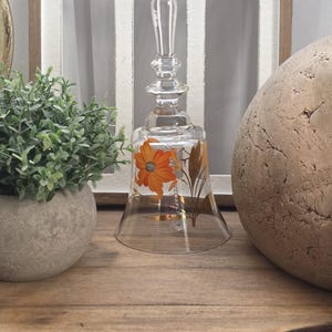 May include: A clear glass bell with an orange floral design. The bell has a long, clear handle and a gold-colored clapper. The bell is sitting on a wooden surface, with a small green plant in a gray pot to the left.