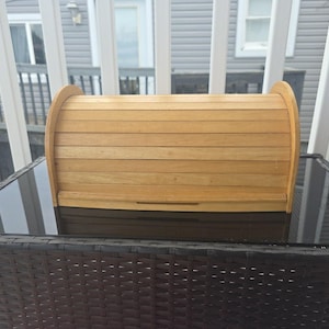 May include: A light brown wooden breadbox with a roll-top lid. The breadbox is made of horizontal wooden slats and has curved sides. It sits on a black, textured surface.