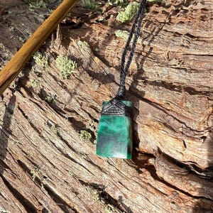 May include: A rectangular green pendant with a black cord necklace. The pendant has a dark green and black marbled appearance. The necklace is displayed on a piece of weathered wood with green grass in the background.