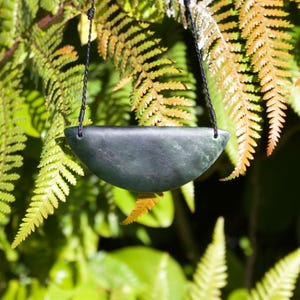 Può includere: Un ciondolo in pietra verde scuro a forma di semicerchio con una finitura liscia e opaca. Il ciondolo presenta due piccoli fori agli angoli superiori, attraverso i quali è infilato un sottile cordino intrecciato nero per formare una collana.