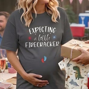 May include: A person wearing a grey t-shirt with the text "Expecting a little firecracker" at a garden party. The shirt has a red, white, and blue design. A friend is giving a gift of a baby onesie.