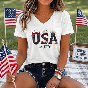 May include: A white V-neck t-shirt featuring the text USA in a dark blue floral pattern with red bows, and the text est. 1776 below it. The shirt is displayed alongside small American flags and a sign reading FOURTH OF JULY CELEBRATION.