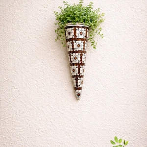 May include: A decorative, cone-shaped wall planter featuring a brown and white floral design, filled with green plants. The planter is mounted on a textured white wall. The design is reminiscent of vintage decor. The planter is approximately 30 cm long.