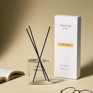 May include: A clear square glass reed diffuser bottle with black reeds and a white rectangular box labeled KODAXTRE Atmos Soft Cedar. The bottle and box both display the text Reed Diffuser 100ml/3.38 fl oz. The set is presented with a book and glasses.