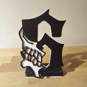 Peut inclure: Une lettre S autoportante avec une finition noire pailletée et un contour blanc. La lettre présente un motif de crâne blanc intégré sur le côté gauche, posée sur une base rectangulaire noire. L'objet a une apparence de police audacieuse.