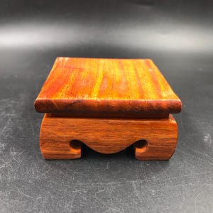 Könnte beinhalten: Kleiner, rechteckiger Holz-Präsentationsständer mit rotbraunem Finish. Die Oberseite ist flach, und die Basis hat ein geschwungenes Design mit dekorativen Ausschnitten. Der Ständer dient wahrscheinlich zur Präsentation kleiner Objekte.