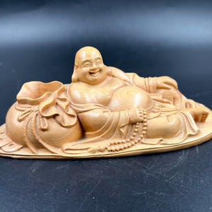 May include: A light brown wooden carving of a reclining, smiling figure with a large belly, resting on a base next to a tied sack. The figure holds a string of prayer beads in one hand and is depicted in a relaxed, lying position.