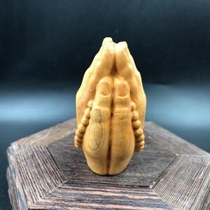 Könnte beinhalten: Eine Holzskulptur von zum Gebet gefalteten Händen. Die Hände sind in einem hellbraunen Farbton geschnitzt, mit detaillierten Fingern und einer strukturierten Oberfläche. Die Skulptur steht auf einem Holzsockel mit dunklem Hintergrund.