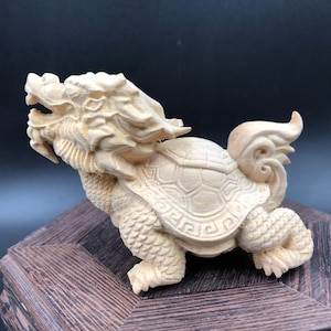 Könnte beinhalten: Detaillierte Holzschnitzerei eines Fabelwesens, einer Mischung aus Drache und Schildkröte. Die Skulptur ist hell, mit komplizierten Details an Kopf, Panzer und Beinen. Die Schnitzerei wird auf einem dunklen Holzsockel präsentiert.