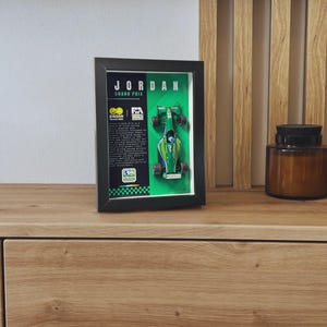 Peut inclure: Un cadre noir de type boîte vitrine contenant une miniature de voiture de course de Formule 1 Jordan Grand Prix verte, montée sur un fond vert avec du texte blanc détaillant l'histoire de l'équipe, incluant le logo Jordan Grand Prix et le logo F1.
