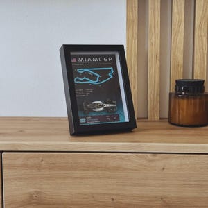 Op de afbeelding: Een zwarte fotolijst met een lichtblauwe omtrek van het Miami GP-circuit, een miniatuur Formule 1-auto en tekst waaronder "MIAMI GP", "Kimi Antonelli" en "Mercedes AMG F1" op een donkere achtergrond met een rasterpatroon.