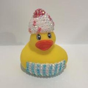 May include: A yellow rubber duck with an orange beak and black eyes. The duck is adorned with a white and pink beaded hat and a blue and white beaded band around its base. The duck is a whimsical decorative item.