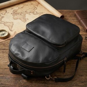 May include: A black leather backpack featuring a large main compartment, a smaller front zippered pocket, and a rectangular leather patch on the front. The bag includes antique brass-toned metal zips, a top handle, and adjustable shoulder straps.