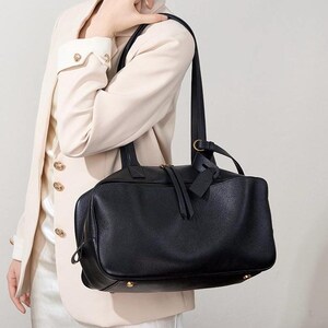 May include: A black leather tote bag with a rectangular shape and a top handle. The bag has a zipper closure and a small tag attached to the handle. The bag is carried over the shoulder.
