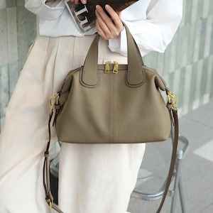 May include: A taupe leather handbag with a top handle and a detachable shoulder strap. The bag features gold-toned hardware, including zippers and clasps. The bag is a neutral color and has a structured shape.