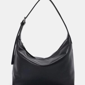May include: Black shoulder bag with a curved design. The bag has a long, adjustable strap and a zipper closure. The bag is made of a smooth, leather-like material and is suitable for everyday use.