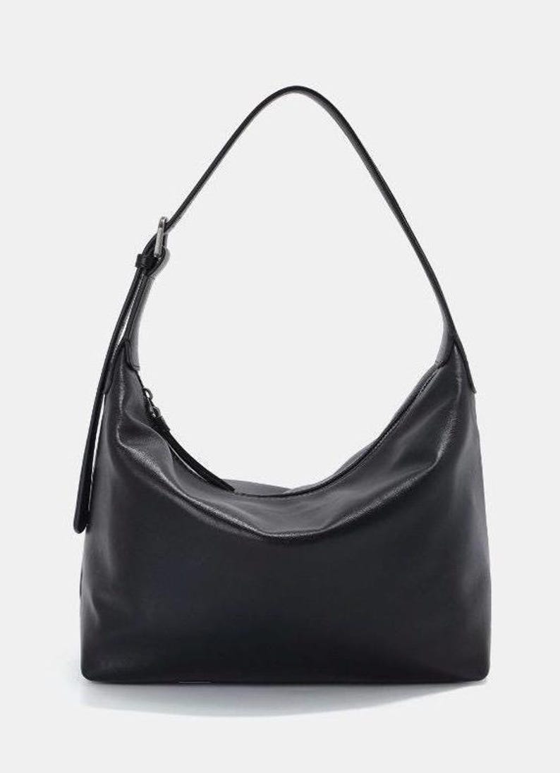 May include: Black shoulder bag with a curved design. The bag has a long, adjustable strap and a zipper closure. The bag is made of a smooth, leather-like material and is suitable for everyday use.