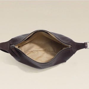 May include: A dark brown leather fanny pack with a zipper closure. The bag is open, revealing a tan interior. The strap features a silver-colored buckle.