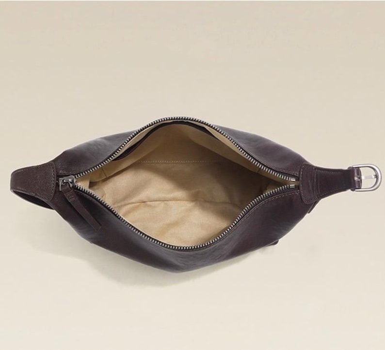 May include: A dark brown leather fanny pack with a zipper closure. The bag is open, revealing a tan interior. The strap features a silver-colored buckle.